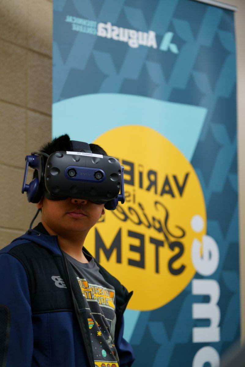 An African American male stands solemnly wearing VR equipment in front of an 奥古斯塔 Technical College VARIETY is Spice STEM banner.